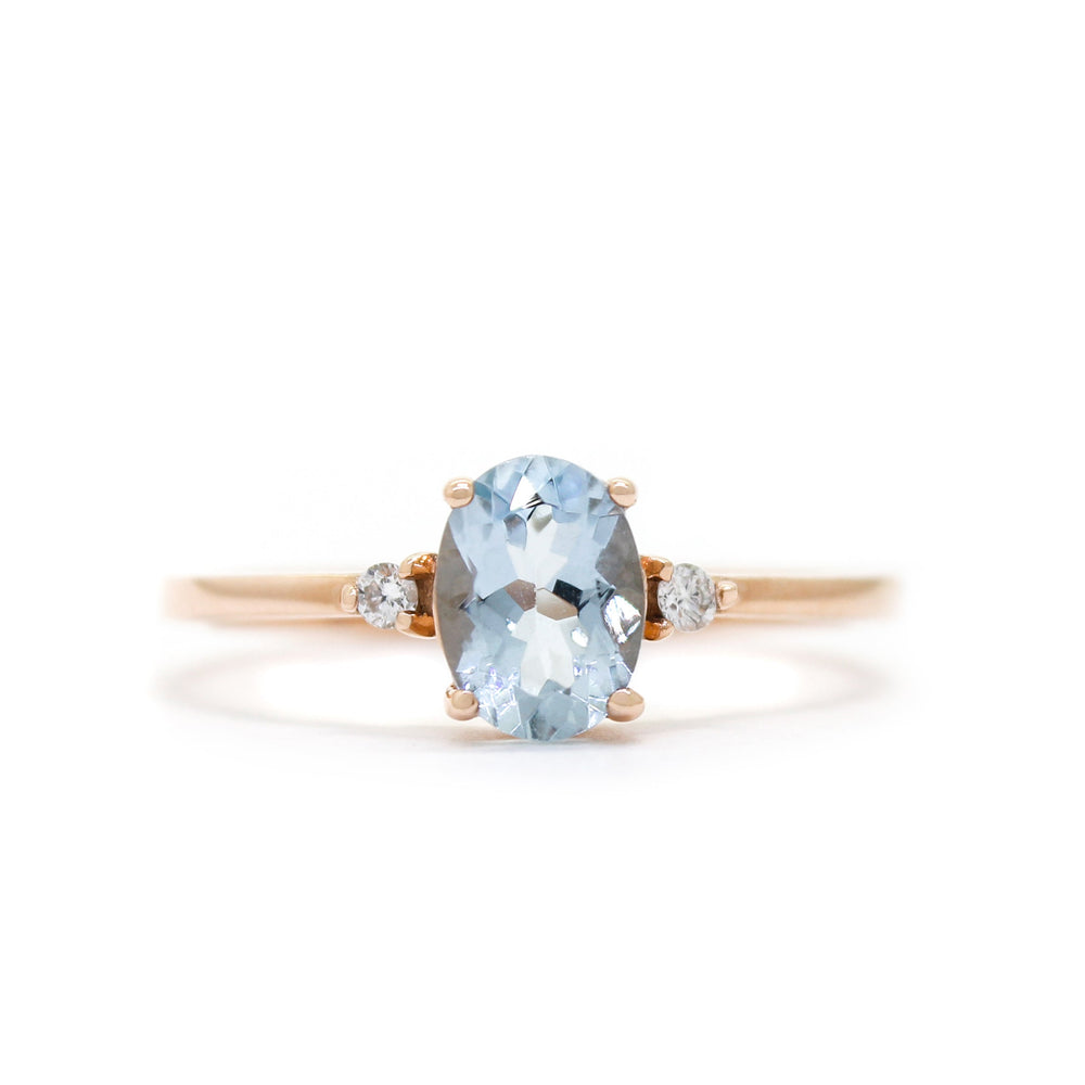 A product photo of a rose gold aquamarine ring sitting against a white background. The rose gold band is plain and smooth, and the centre oval-cut light blue aquamarine stone is framed by a single white diamond on either side.