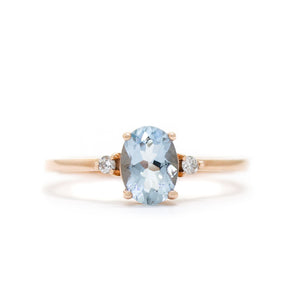 A product photo of a rose gold aquamarine ring sitting against a white background. The rose gold band is plain and smooth, and the centre oval-cut light blue aquamarine stone is framed by a single white diamond on either side.