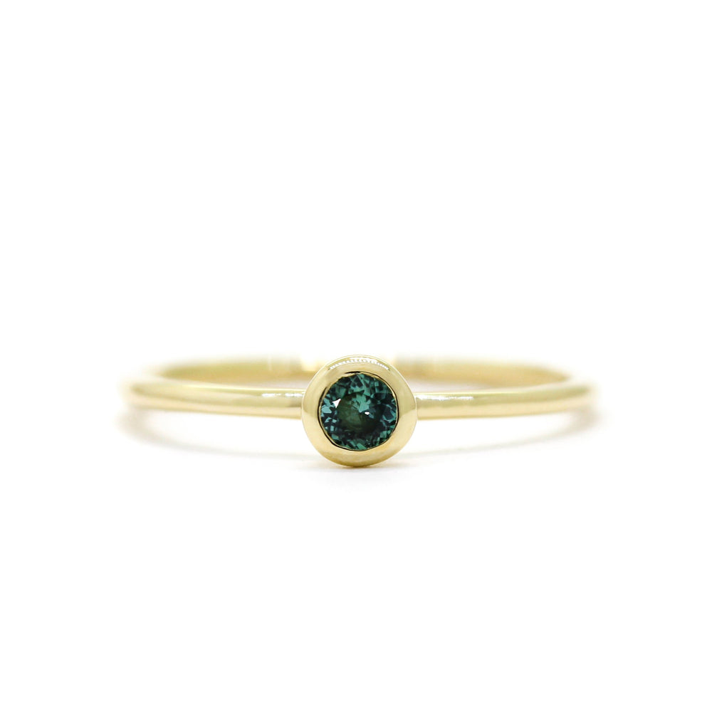 A product photo of a delicate yellow gold stacking ring with a tiny, bezel-set green tourmaline in the centre sitting on a white background. The band is slim and thread-like, with the focus drawn to the petite 3mm glinting green centre stone.