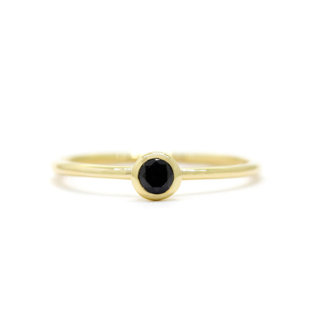 A product photo of a delicate yellow gold stacking ring with a tiny, bezel-set black diamond in the centre sitting on a white background. The band is slim and thread-like, with the focus drawn to the petite 3mm glinting black centre stone.