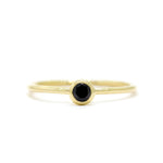 A product photo of a delicate yellow gold stacking ring with a tiny, bezel-set black diamond in the centre sitting on a white background. The band is slim and thread-like, with the focus drawn to the petite 3mm glinting black centre stone.