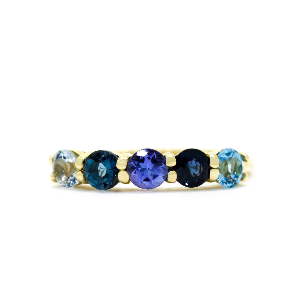 A product photo of a bold, multi-gemstone ring in 9k yellow gold – made up of 5 blue coloured jewels – sitting on a white background. From left to right, 4.5mm round-cut aquamarine, blue tourmaline, tanzanite, sapphire and blue topaz stones make up a bold statement piece.