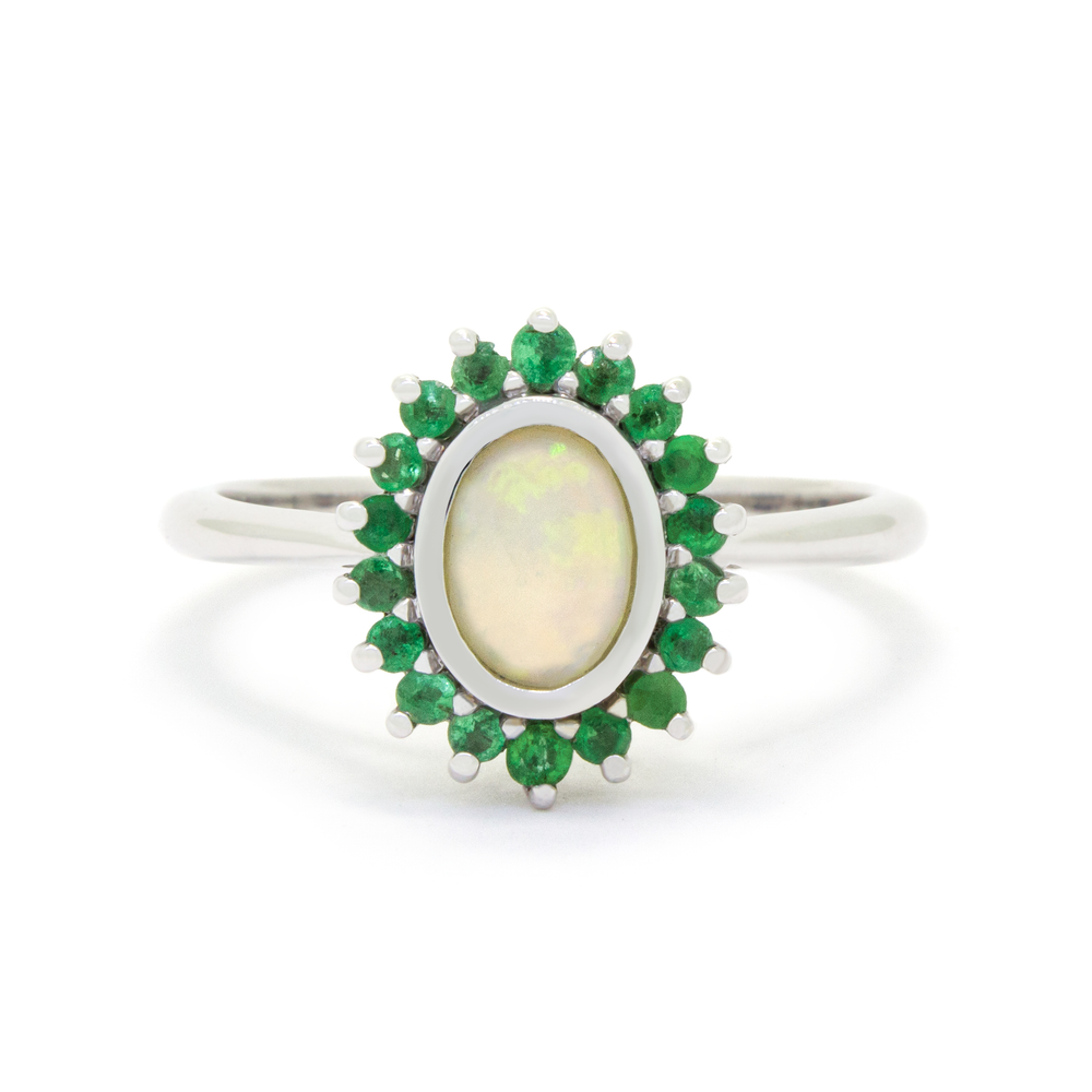 A product photo of a vintage styled white opal ring in 9k white gold on a white background. A large, fiery oval stone in the centre reflects multi-coloured light, and is framed by a ring of green emeralds. The band is simple and rounded.