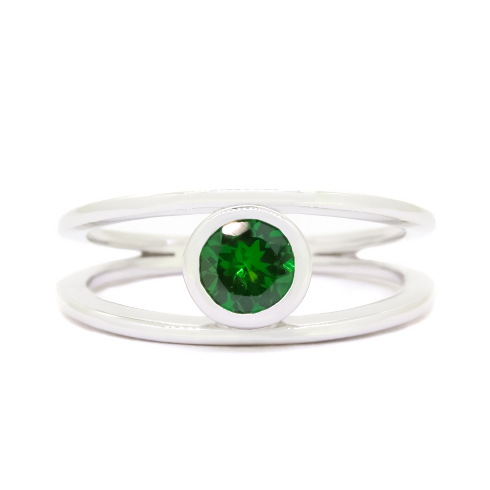 A product photo of a split-band white gold ring with a bezel-set tsavorite centre stone sitting on a white background. The band splits at the base of the ring, separating to meet at the top and bottom of the verdant-green tsavorite centre stone, framed in a thick layer of gold, holding it in place.  The green gemstone colour be a good emerald substitute.