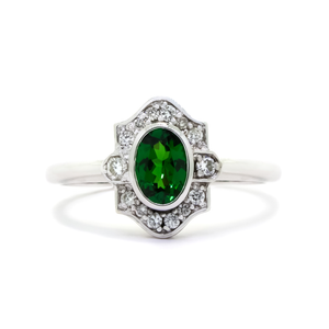 A product photo of a vintage-styled tsavorite and diamond ring in 9k white gold on a white background. The oval-shaped tsavorite is held in place by a bezel setting, and is surrounded by an ornate white gold frame embedded with diamonds, reminiscent of vintage jewellery styles of the past. The green gemstone colour be a good emerald substitute.