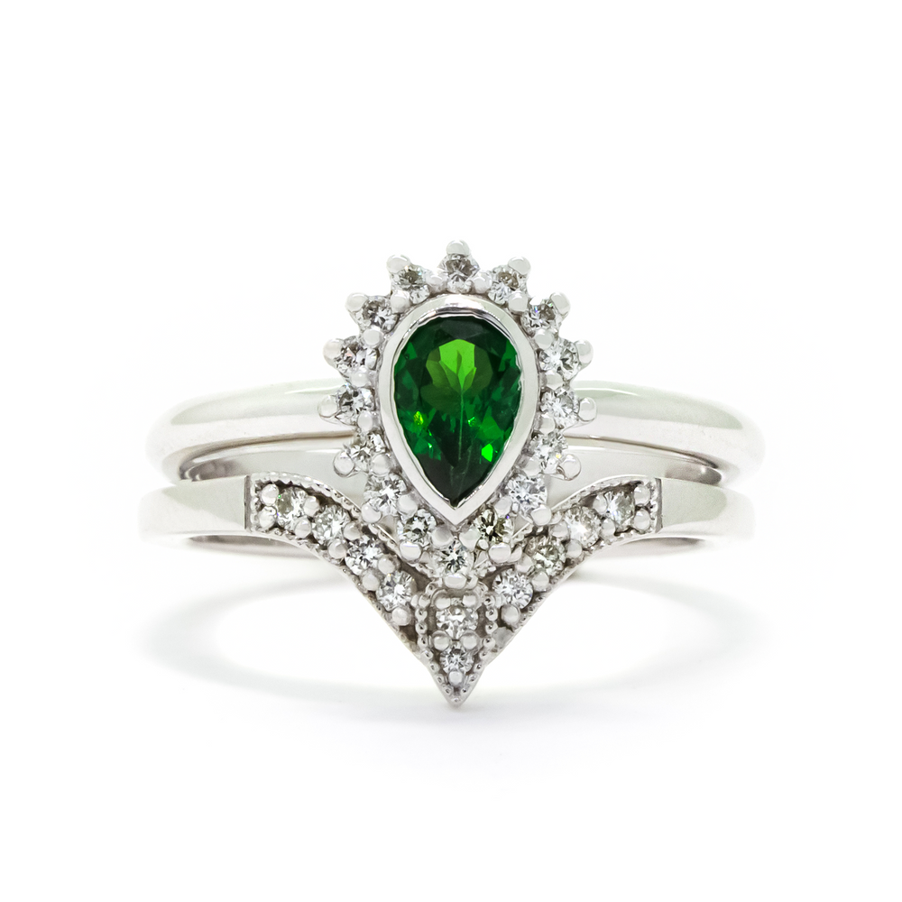 A product photo of a bridal/wedding set made of solid 9k white gold. The engagement ring is made up of a pear-shaped tsavorite gemstone, framed by a halo of diamonds and elegant gold detailing. The band is rounded and smooth, making for comfortable wear. The wedding band is designed to match the engagement ring perfectly, with 10 diamonds embedded along its length to echo the halo that frames the engagement ring's stone. The green gemstone colour be a good emerald alternative for wearers born in May.