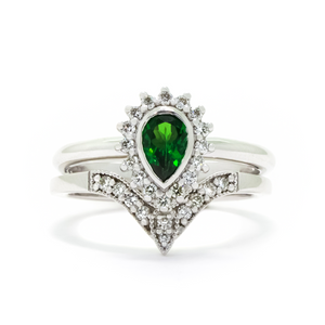 A product photo of a bridal/wedding set made of solid 9k white gold. The engagement ring is made up of a pear-shaped tsavorite gemstone, framed by a halo of diamonds and elegant gold detailing. The band is rounded and smooth, making for comfortable wear. The wedding band is designed to match the engagement ring perfectly, with 10 diamonds embedded along its length to echo the halo that frames the engagement ring's stone. The green gemstone colour be a good emerald alternative for wearers born in May.
