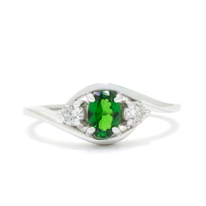 A product photo of a mystical tsavorite and diamond fantasy ring in solid 9k white gold on a white background. The deep, verdant-green tsavorite is held in place by 4 claws and reflects light off of its many facets, while the diamonds on either side are held by 3 each. The smooth white gold band curves elegantly, meeting at the top and bottom of the tsavorite and diamond arrangement. The green gemstone colour be a good emerald substitute.
