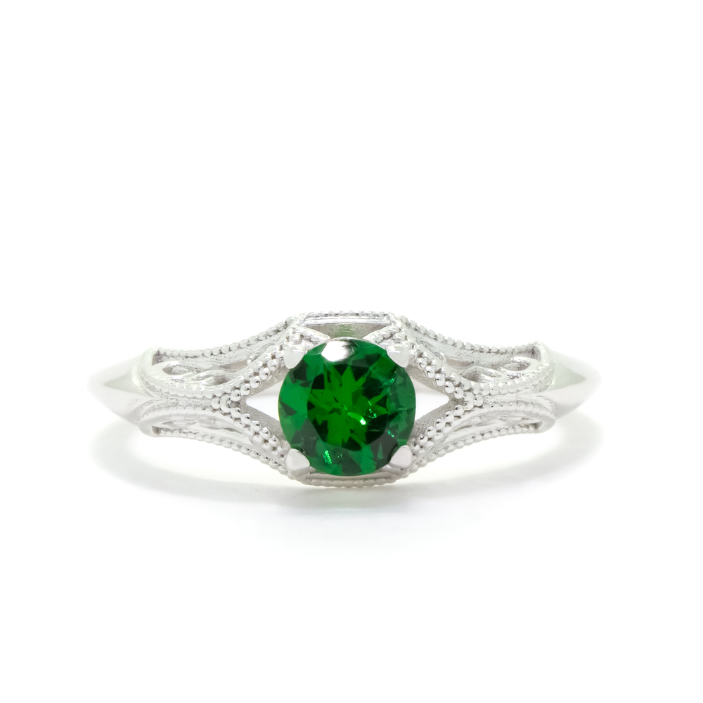 A product photo of an ornate, filigree-detailed white gold ring - reminiscent of an elvish fantasy ring with a deep forest green tsavorite centre stone. The band is detailed with delicate curves and patterns for about half of its length around the dazzling tsavorite stone, before smoothing out towards the back of the ring. The green gemstone colour be a good emerald substitute.