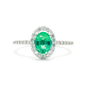 A product photo of a fine emerald ring in 14k white gold on a white background. A large, brilliant oval jewel in the centre reflects mint-coloured light, and is framed by a ring of white diamonds. A simple round band continues the diamond detailing for half of its length, smoothing out into a simple white gold back.