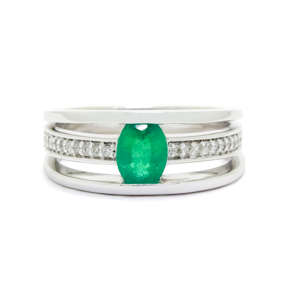 A product photo of a white gold trio-banded ring with diamond detailing and a natural emerald centre stone sitting on a white background. The top and bottom bands are smooth and minimalistic, hugging the top and bottom of the oval emerald centre stone, whilst the middle band (meeting on either side of the stone) is bejewelled with many tiny diamonds.