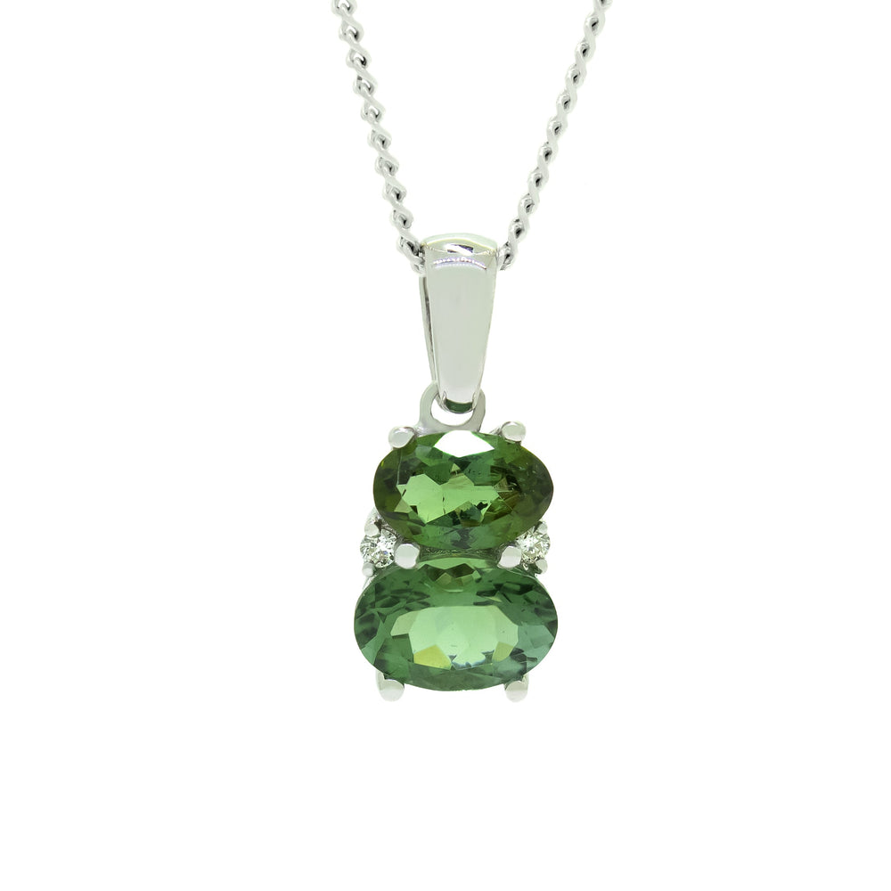 A product photo of a green tourmaline and diamond pendant in 9k white gold sitting on a white background. The pendant is made up of two horizontal oval-shaped green tourmaline stones, the bottom one being 7x5mm and the top being 6x4mm. The spaces between the two stones are filled with one small diamond on either side.