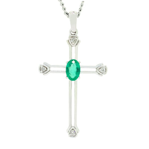 A product photo of a white gold emerald and diamond pendant in the shape of a Christian cross. At the ends of each limb, a tiny white diamond marks the tip, to a total of 4 diamonds. At the heart of the cross is a dainty fine emerald centre stone.