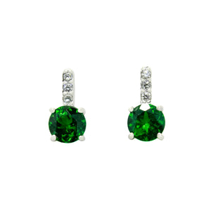 A product photo of a pair of white gold tsavorite earrings sitting against a white background. The simple circle-cut stones are contrasted by the white diamond trio protruding in a straight line above each stone, encased in white gold.