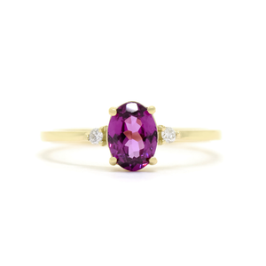 A product photo of a yellow gold grape garnet ring sitting against a white background. The gold band is plain and smooth, and the centre oval-cut warm purple garnet stone is framed by a single white diamond on either side.