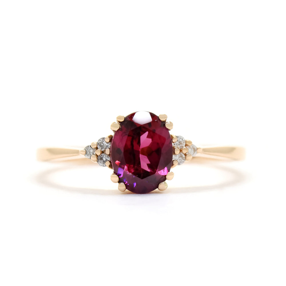 A product photo of a rose gold rhodalite and diamond trio ring sitting on a white background. The oval purple rhodalite gemstone stands in stark contrast to the little clusters of three white diamond stones on either side.