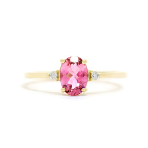A product photo of a yellow gold pink tourmaline ring sitting against a white background. The gold band is plain and smooth, and the centre oval-cut bright pink tourmaline stone is framed by a single white diamond on either side.