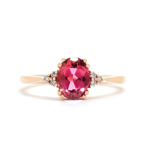 A product photo of a rose gold pink tourmaline and diamond trio ring sitting on a white background. The oval magenta pink tourmaline gemstone stands in stark contrast to the little clusters of three white diamond stones on either side.