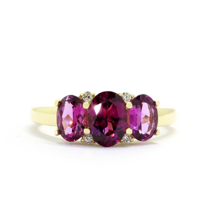 A product photo of a Graoe Garnet Trio & Diamond Ring in Yellow Gold sitting on a plain white background. The largest garnet centrestone measures 7x5mm, with 4 tiny diamonds nestled against it, two on each side. The grape garnet stones are deep and warm, reflecting magenta hues across their multi-faceted edges.