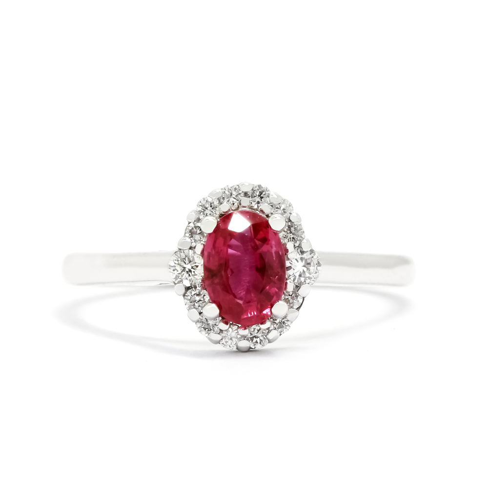 A product photo of a white gold ruby engagement ring sitting on a white background. A flowery halo of diamond and white gold detailing frames the 6x4mm oval centre stone, starkly contrasting the bright white of the diamonds to the dazzling colour reflections of the fine ruby gemstone. The deep magenta ruby jewel reflects brightly from its many edges.