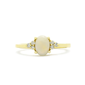 A product photo of a yellow gold white opal and diamond trio ring sitting on a white background. The oval opalite gemstone stands in stark contrast to the little clusters of three white diamond stones on either side.