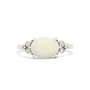 A product photo of a white gold white opal and diamond trio ring sitting on a white background. The large oval opalite gemstone stands in stark contrast to the little clusters of three white diamond stones on either side.