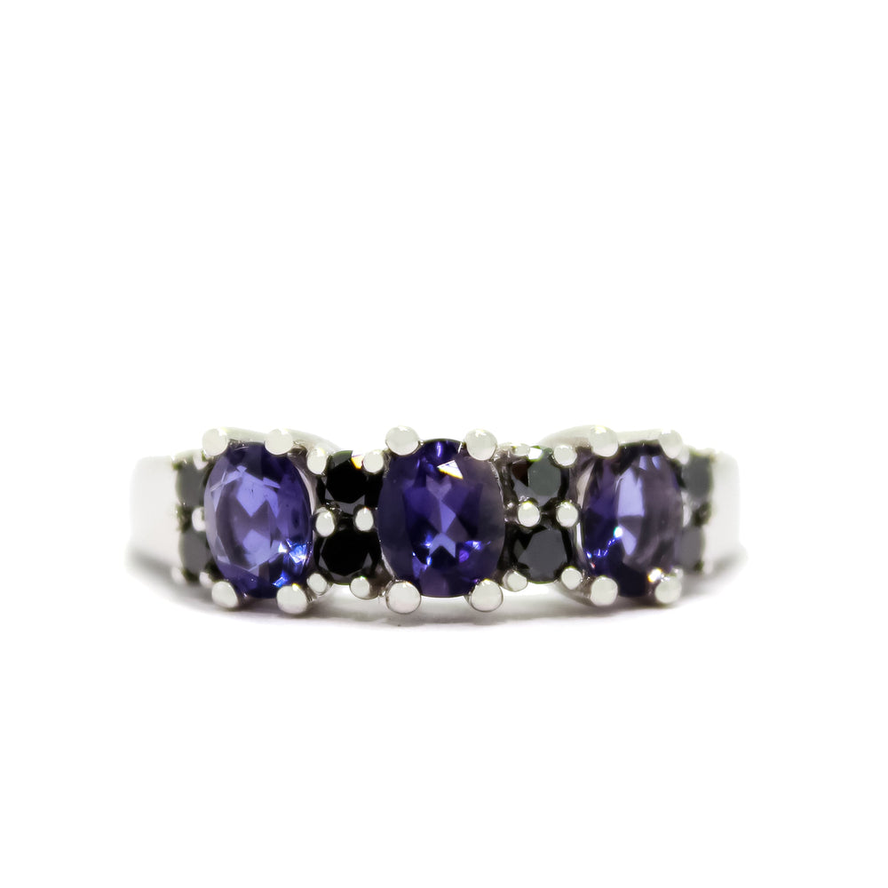 A product photo of a bold and lavish iolite and black diamond ring in 9k white gold. The ring is made up of three 5x4mm oval iolite stones, while black 2 diamonds fill the spaces between and on either side of the iolites - to a total of 0.32ct for 8 diamonds. The deep blue colour could make it a good sapphire or tanzanite alternative.
