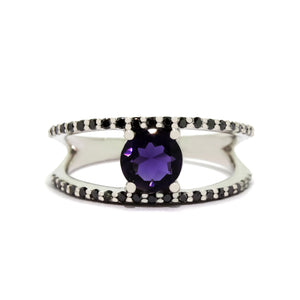 A product photo of an iolite and black diamond ring in solid 9 karat white gold sitting on a white background. The large iolite gemstone is held in place by 4 claws. The white gold band splits halfway along its length into two smaller bands detailed with black diamonds, and hugs the centre stone's top and bottom. The deep blue colour could make it a good sapphire or tanzanite alternative.