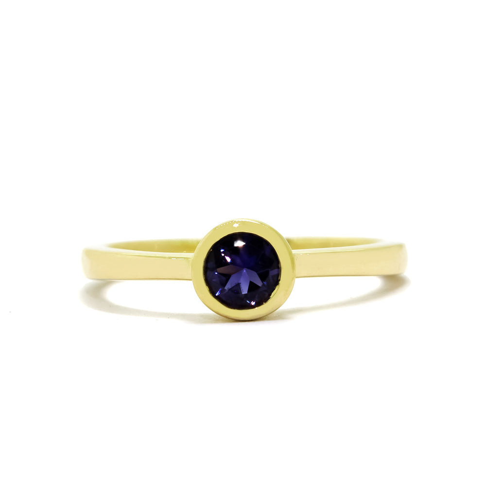 A product photo of a solid 9 karat yellow gold bezel-set solitaire ring. A single, midnight blue iolite gemstone sits encased in a thick layer of golden framing. The dark blue colour could make it a good sapphire or tanzanite alternative.