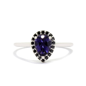 A product photo of an iolite ring with a halo of black diamonds set in solid 9 karat white gold sitting on a white background. A flowery halo of black diamond and white gold detailing frames the large, pear-shaped centre stone. The deep blue iolite reflects violet blue and indigo colours from its many edge, and could serve as an affordable alternative to sapphire or tanzanite.