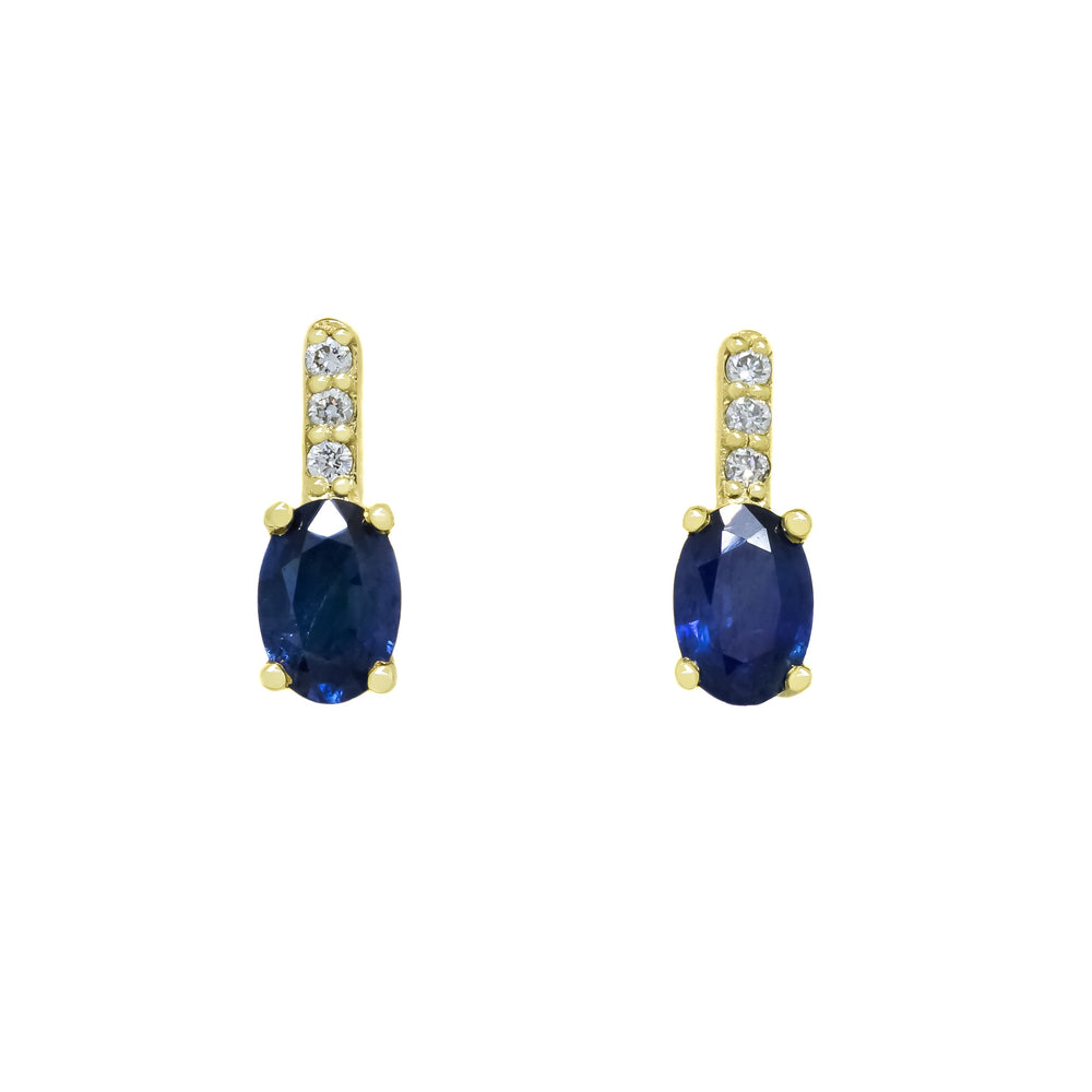 A product photo of a pair of yellow gold sapphire earrings sitting against a white background. The simple oval-cut stones are a shade of saturated, cold blue - contrasted by the white diamond trio protruding in a straight line above each midnight-coloured stone, encased in yellow gold.