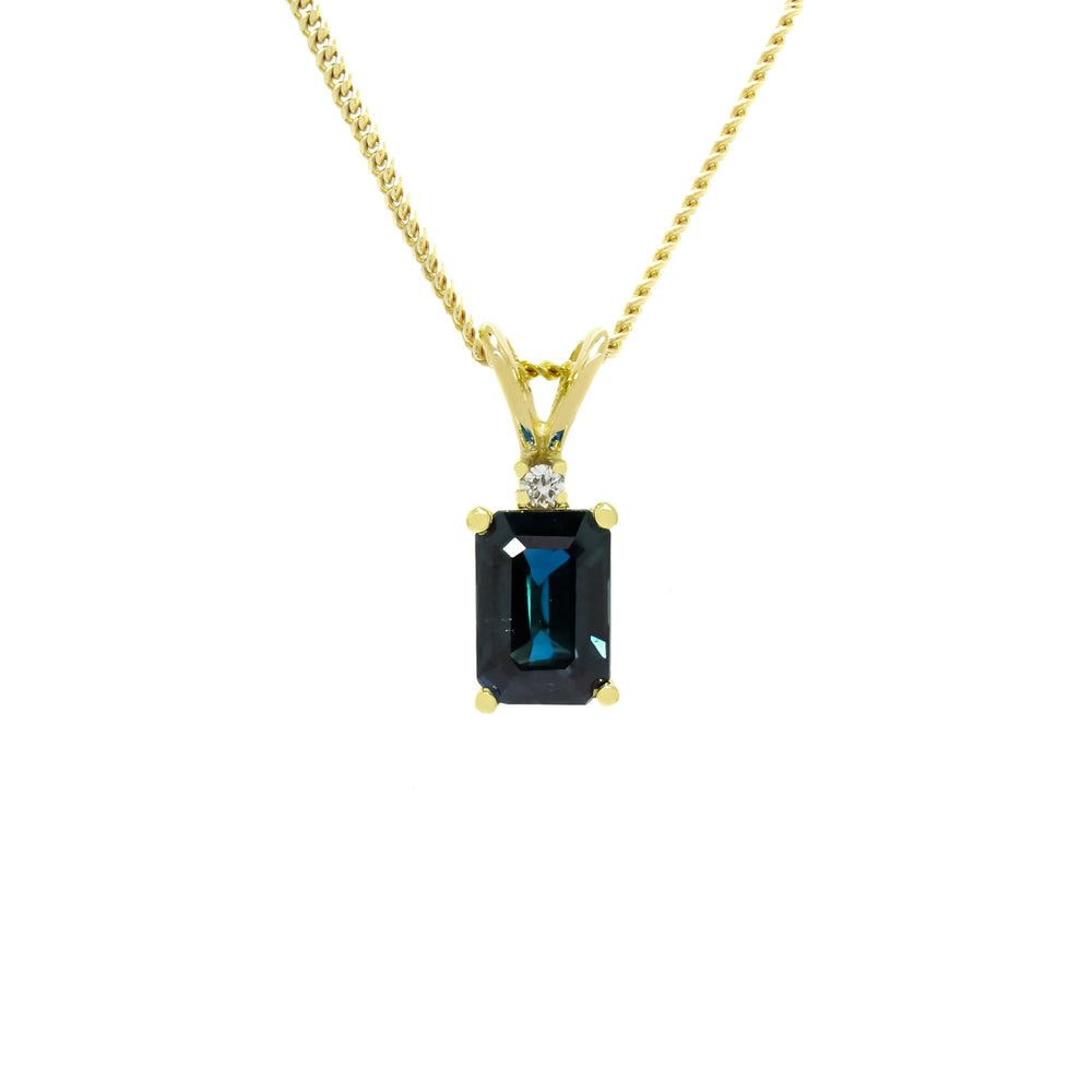 A product photo of a rectangular sapphire and diamond pendant in 9k yellow gold suspended against a white background. The rectangular, emerald-cut sapphire is a stunningly deep ocean blue colour. A single white diamond sits atop it, before meeting the split golden bail.
