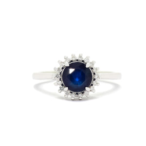
            
                Load image into Gallery viewer, A product photo of a white gold sapphire ring sitting on a white background. A flowery halo of diamond and white gold detailing frames the large, round centre stone. The deep blue sapphire reflects blue and azure colours from its many edges.
            
        