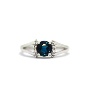 A product photo of a sophisticated blue sapphire split-band ring with diamond framing on a white background. 4 diamonds sit on either side of the deep midnight blue sapphire gemstone, with the split band on either side holding the jewel and diamond arrangement in place.