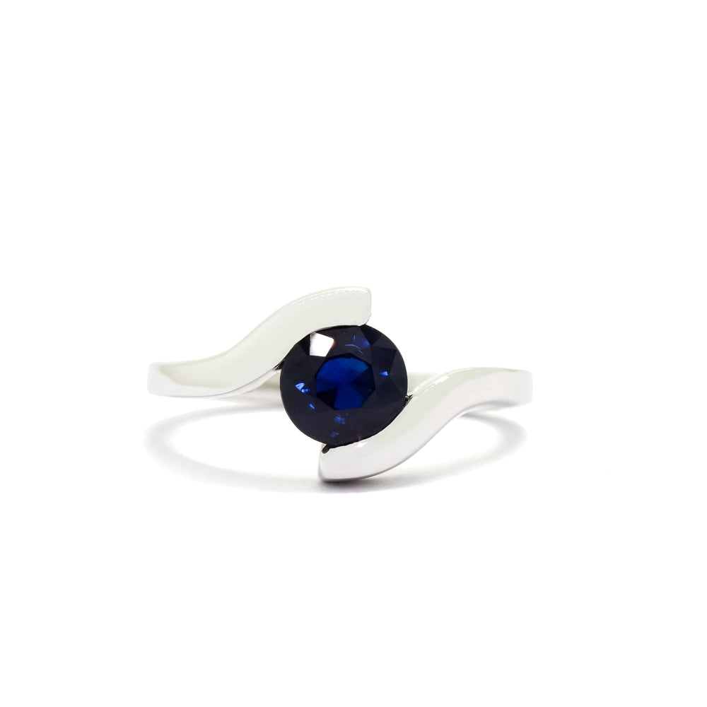 A product photo of a white gold sapphire ring sitting on a white background. The band splits and curves around the single, round sapphire centre stone, meeting at its top and bottom to hold it in place.