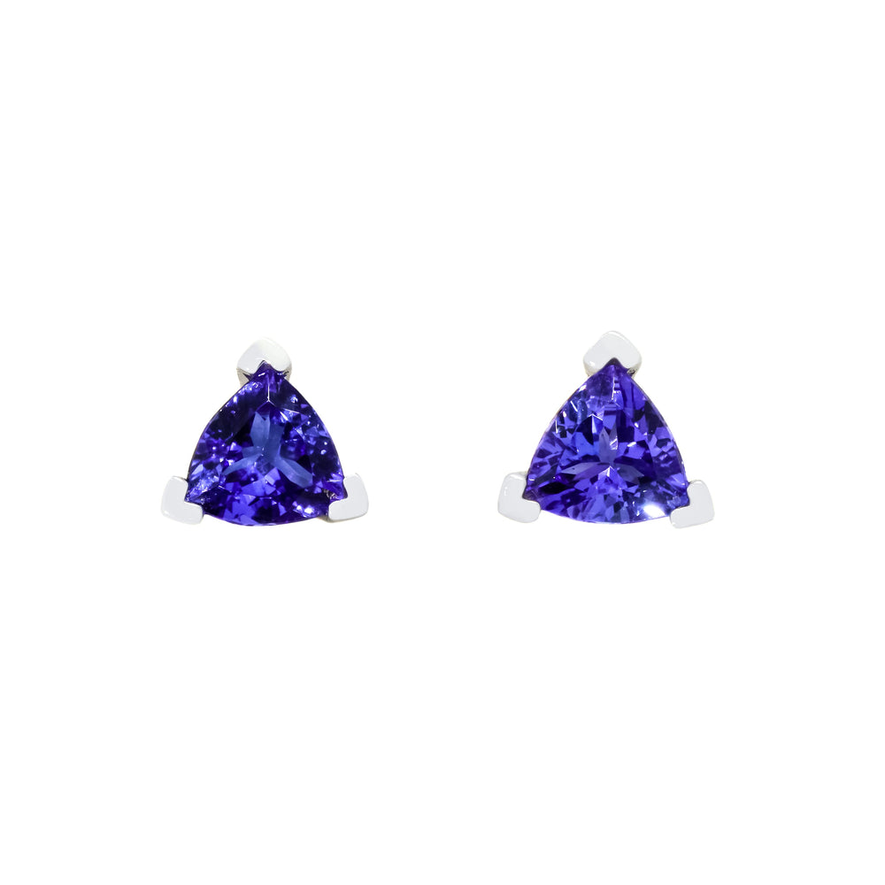 A product photo of two white gold stud earrings sitting on a white background. Held in place by 3 silver claws each are two dazzling 7mm trilliant-cut indigo blue tanzanite gemstones, reflecting shades of cool violet from their many edges.