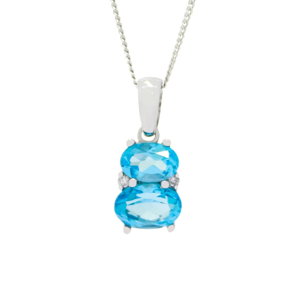 A product photo of a blue topaz and diamond necklace in 9k white gold sitting on a white background. The pendant is made up of two horizontal oval-shaped blue topaz stones, the bottom one being 7x5mm and the top being 6x4mm. The spaces between the two stones are filled with one small diamond on either side.