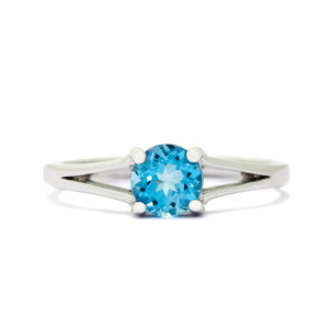 A product photo of a white gold split-band blue topaz ring sitting on a white background. The ring is simple, yet sophisticated in its elegance - splitting halfway along the band's length into two golden prongs that meet on either side of the bright, sky-blue centre jewel.