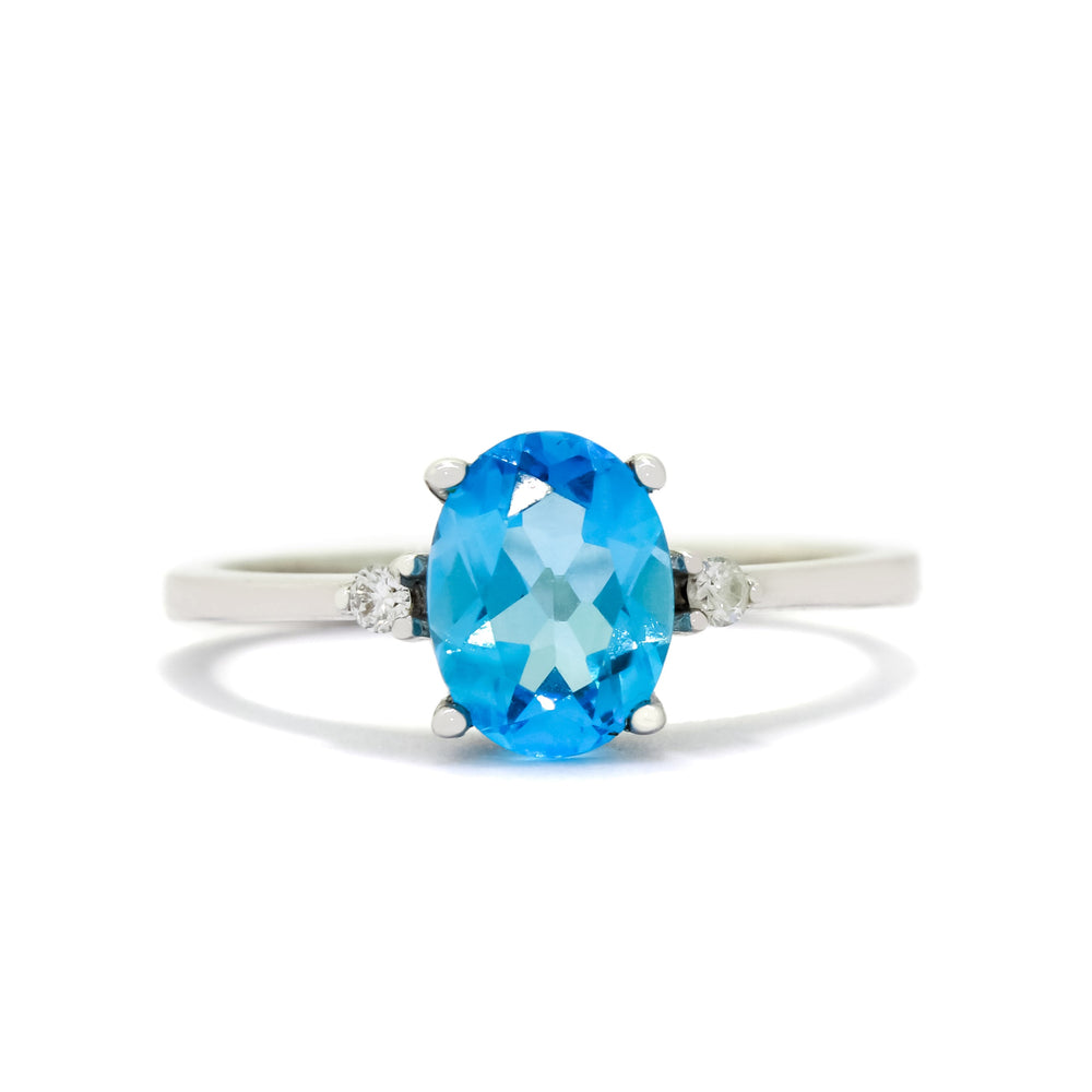
            
                Load image into Gallery viewer, A product photo of a white gold blue topaz and diamond ring sitting against a white background. The white gold band is plain and smooth, and the centre oval-cut light blue topaz stone is framed by a single white diamond on either side.
            
        