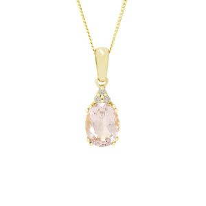 A product photo of a yellow gold pink morganite necklace sitting against a white background. The light pink oval-cut stone is held in place by 4 pairs of claws at its top and bottom, with a trio of white diamonds connecting the stone to the rest of the pendant. It is suspended by a simple yellow gold chain.