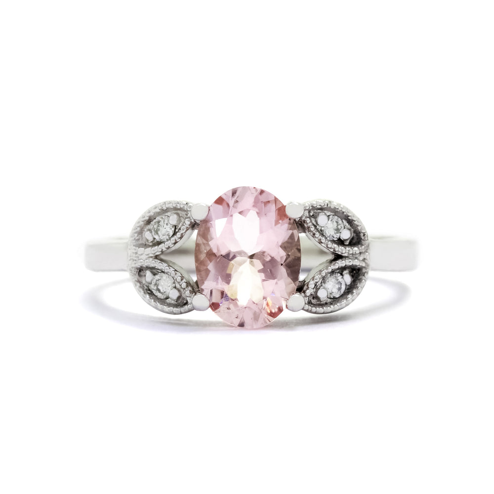 A product photo of an oval morganite and diamond ring with floral gold detailing set in solid 9 karat white gold. The bold 8x6mm pale pink morganite gemstone is held in place by two petal shapes in filigree gold detailing on either side, each "petal" adorned with a single diamond in the centre, to a total of 4.