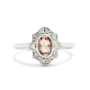 A product photo of a vintage-styled morganite and diamond ring in 9k white gold on a white background. The oval-shaped morganite is held in place by a bezel setting, and is surrounded by an ornate white gold frame embedded with diamonds, reminiscent of vintage jewellery styles of the past.