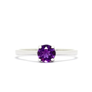 A product photo of a Round Amethyst Solitaire Ring in 9k White Gold sitting on a plain white background. The amethyst centrestone measures 5.5mm across. The stone is a deep purple, and reflects violet and pink hues light across its many faceted edges.