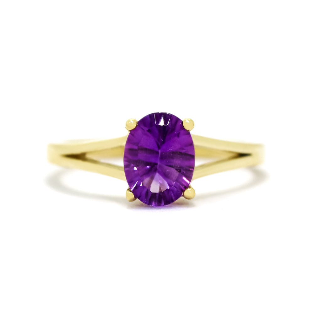 A product photo of a yellow gold split band amethyst ring sitting on a white background. The ring is simple, yet sophisticated in its elegance - splitting halfway along the band's length into two golden prongs that meet on either side of the deep, warm purple centre jewel.