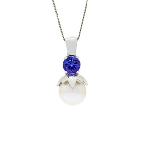 This product image is a close-up of a beautiful solid 9 karat white gold pearl and gemstone pendant design suspended by a golden chain. The pendant primarily features a round-cut tanzanite stone in the centre, set with delicate gold claws above a single white round-shaped pearl. The pearl is held in place by a floral-type setting extending downwards from the tanzanite.