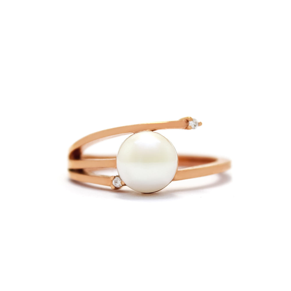 This product image shows an interesting asymmetrical pearl and diamond ring set in solid 9 karat rose gold. In the centre of the design is a single white rounded pearl. The band asymmetrically splits into three slim prongs on the one side of the pearl, with the one meeting the pearl in the middle, while the bottom prong ends short of the ring, with a diamond set between the pearl and the prong. The top prong extends slightly past the edge of the pearl, and is also set with a diamond.