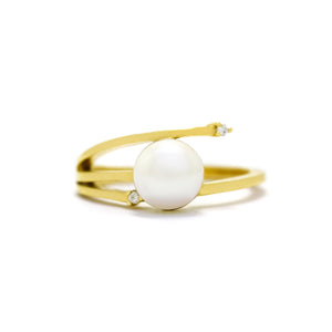 This product image shows an interesting asymmetrical pearl and diamond ring set in solid 9 karat yellow gold. In the centre of the design is a single white rounded pearl. The band asymmetrically splits into three slim prongs on the one side of the pearl, with the one meeting the pearl in the middle, while the bottom prong ends short of the ring, with a diamond set between the pearl and the prong. The top prong extends slightly past the edge of the pearl, and is also set with a diamond.