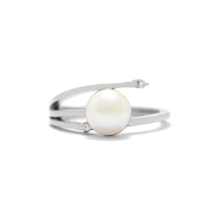 This product image shows an interesting asymmetrical pearl and diamond ring set in solid 9 karat white gold. In the centre of the design is a single white rounded pearl. The band asymmetrically splits into three slim prongs on the one side of the pearl, with the one meeting the pearl in the middle, while the bottom prong ends short of the ring, with a diamond set between the pearl and the prong. The top prong extends slightly past the edge of the pearl, and is also set with a diamond.