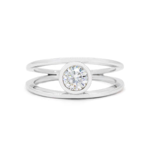 A product photo of a split-band silver ring with a bezel-set white moissanite centre stone sitting on a white background. The silver band splits at the base of the ring, separating to meet at the top and bottom of the dazzling round-cut centre stone, framed in a thick layer of silver, holding it in place.