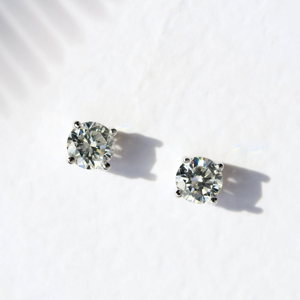 A product photo of a pair of 9 karat white gold lab diamond stud earrings sitting in the sun on a white textured background. The brilliant, colourless half pointer round diamonds measure 5mm across, and are held in place by a simple 4-claw setting.